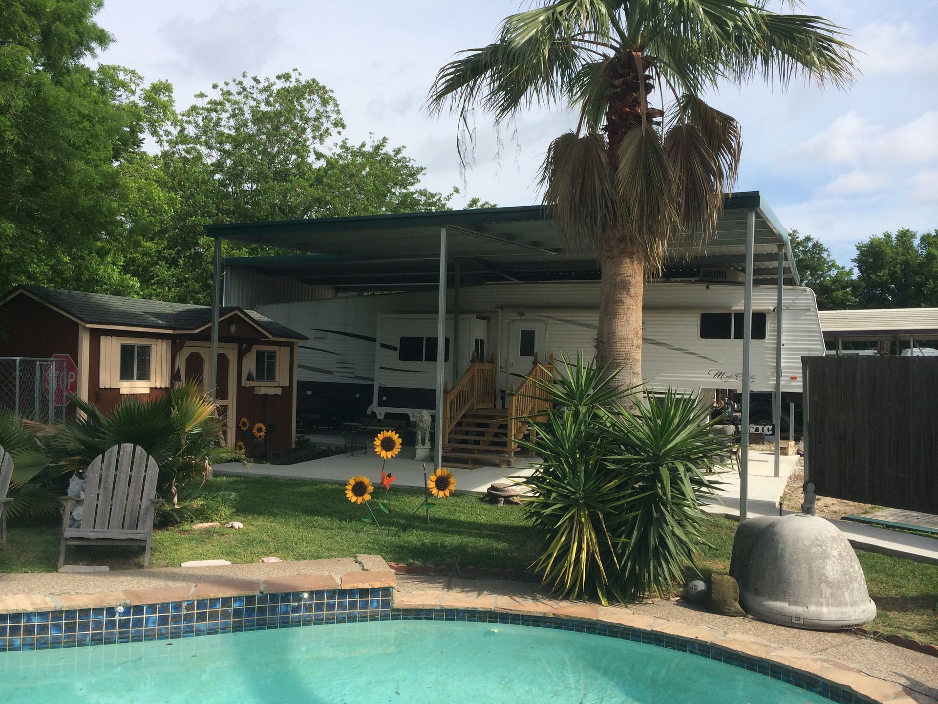 RV under a carport next to a pool and a small shed in a yard with a palm tree.