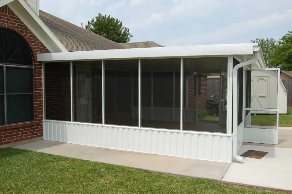 Screened-in patio extension attached to a brick house, featuring white walls, screens, and a door, on a concrete patio.