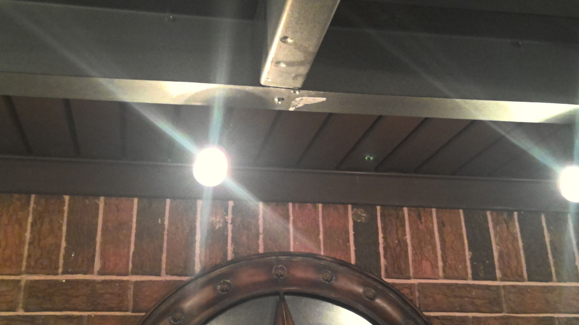 Brick wall with a dark-framed mirror below a ceiling of wooden planks and metal beams with light fixtures.