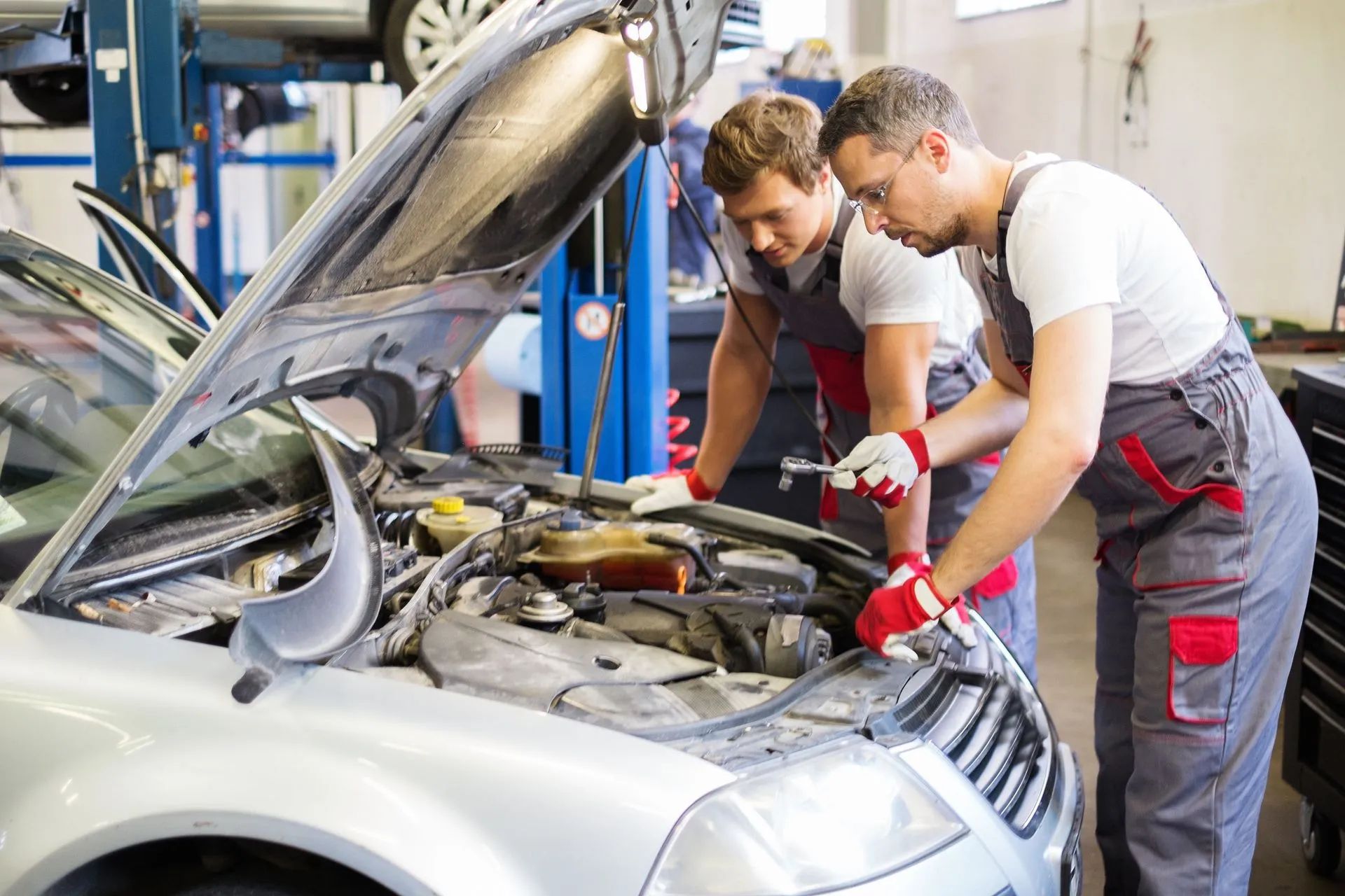 Two mechanics inspect a silver car’s engine with the hood open in a garage