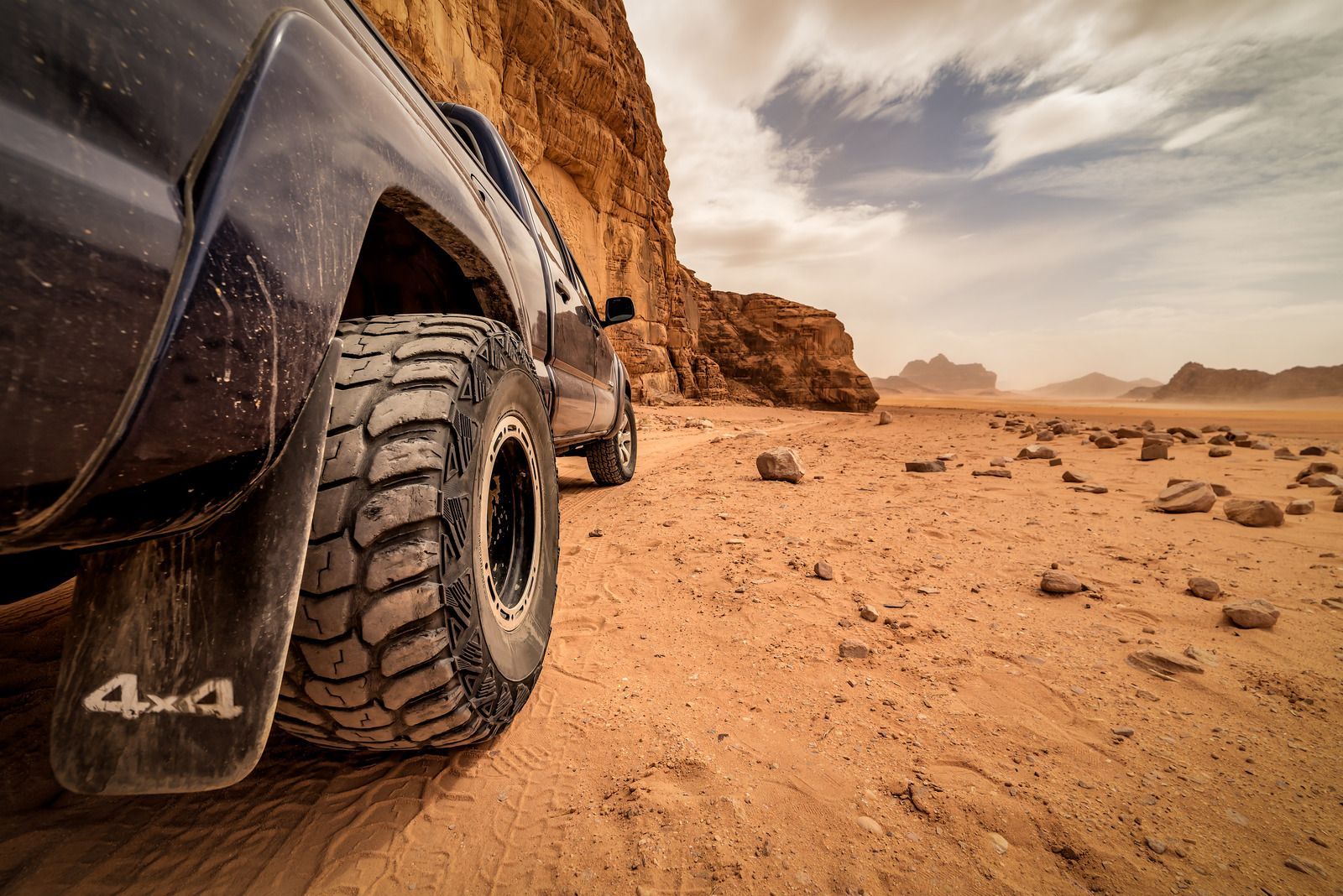 Close-up of a black 4x4 truck driving through a sandy desert beside rocky cliffs