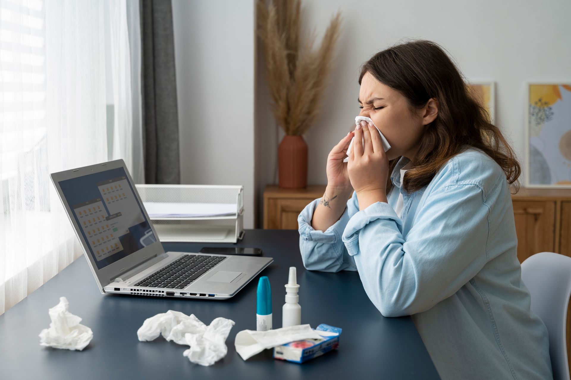 A woman sits at her desk with her laptop open. She is wiping her nose after administering allergy shots.