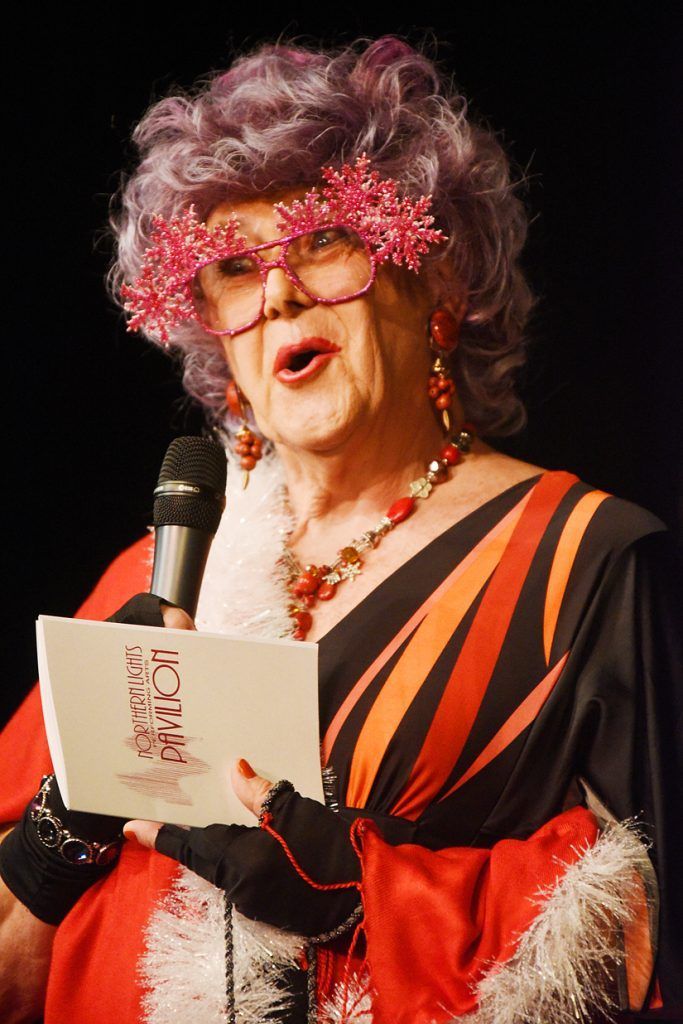 A woman with purple hair and pink glasses is holding a microphone