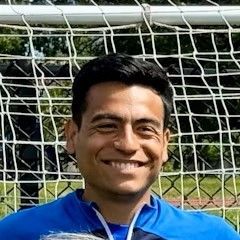 A man in a blue shirt is smiling in front of a soccer goal.