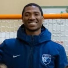 A man in a blue jacket is smiling in front of a soccer goal.