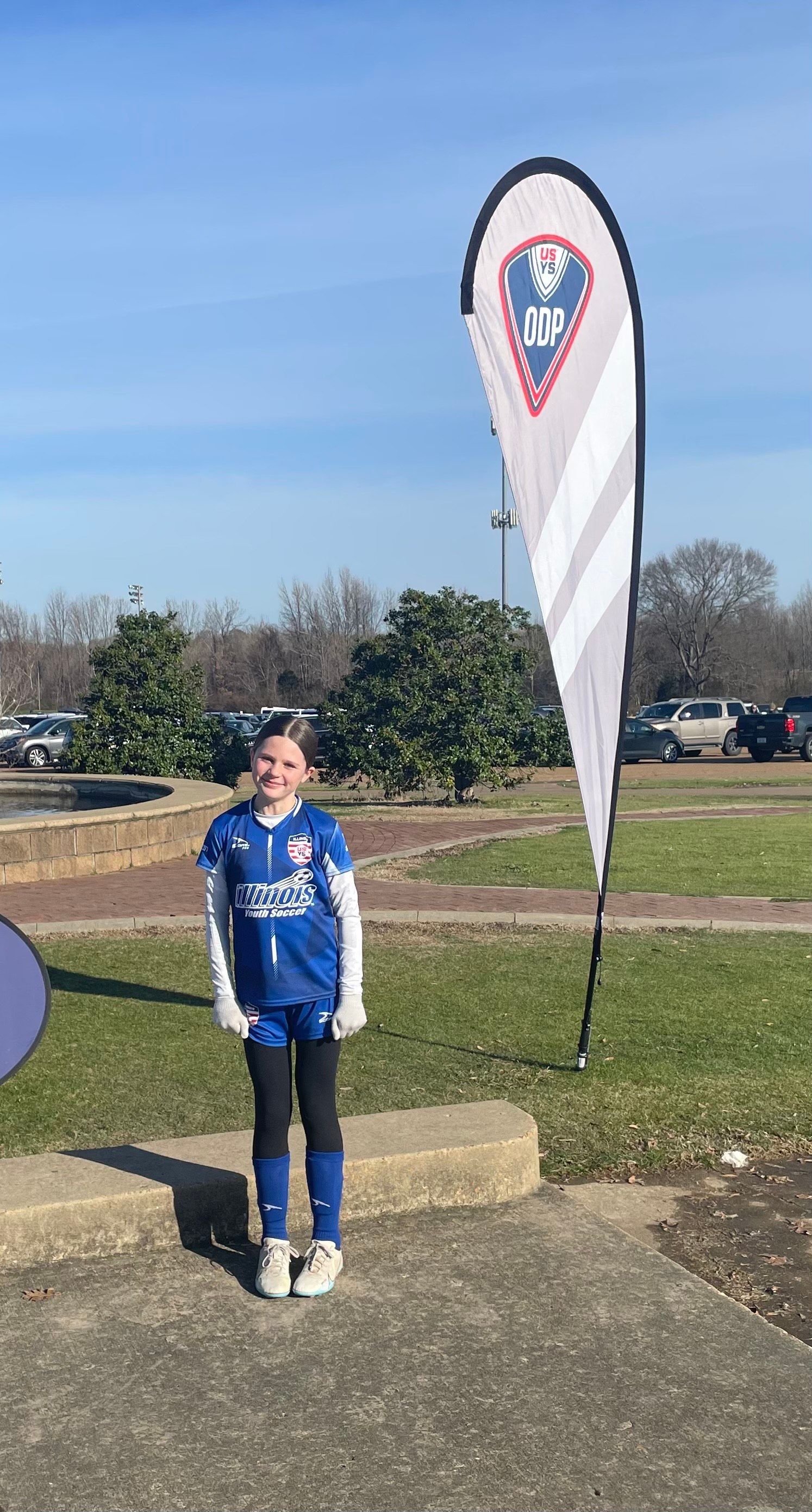 A young girl is standing in front of a flag in a park.