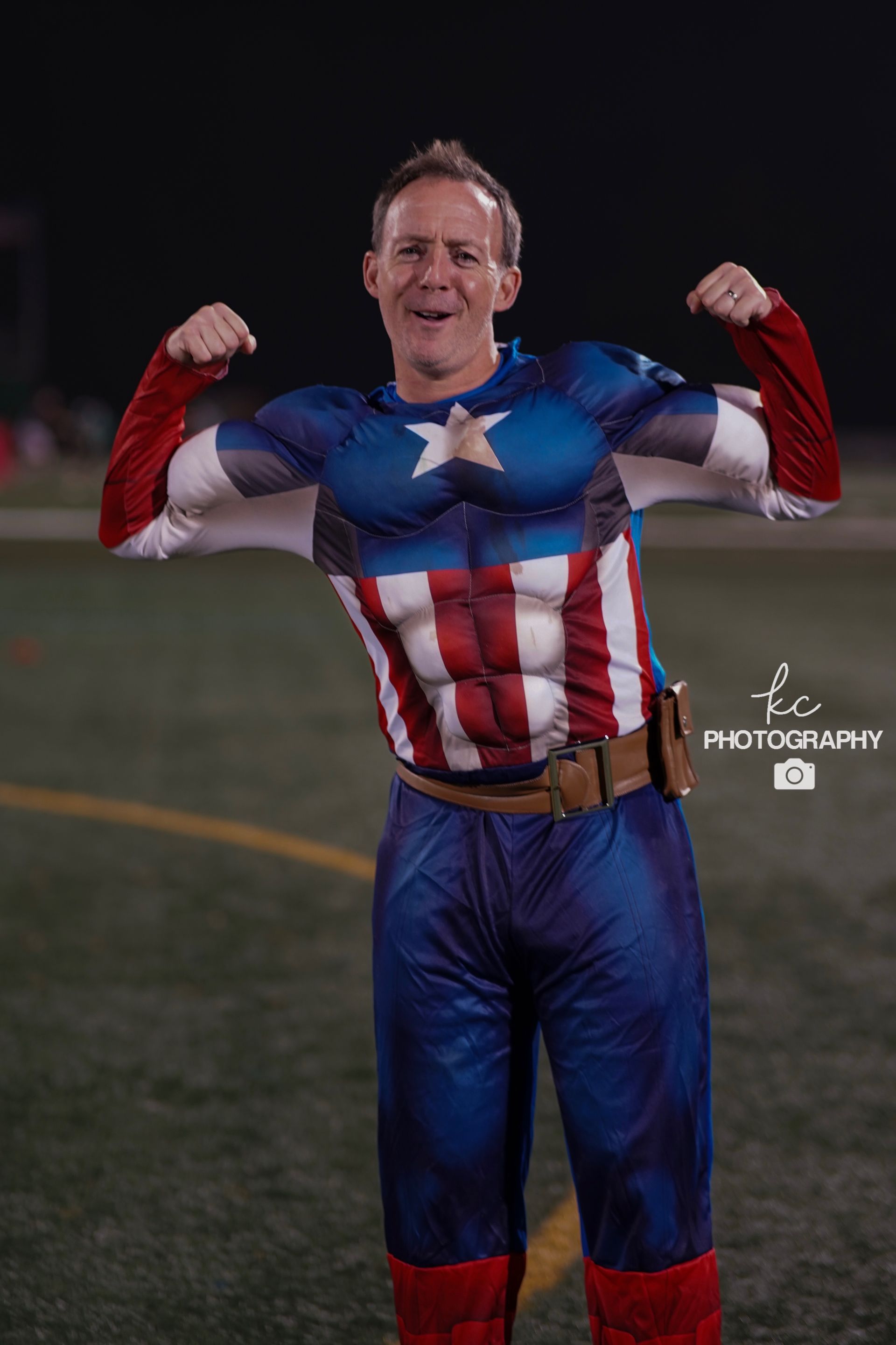 A man in a captain america costume is flexing his muscles on a field.
