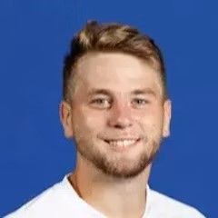 A young man with a beard is smiling for the camera while wearing a white shirt.