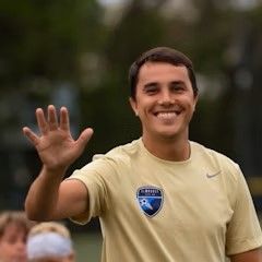 A man in a tan shirt is smiling and waving at the camera.