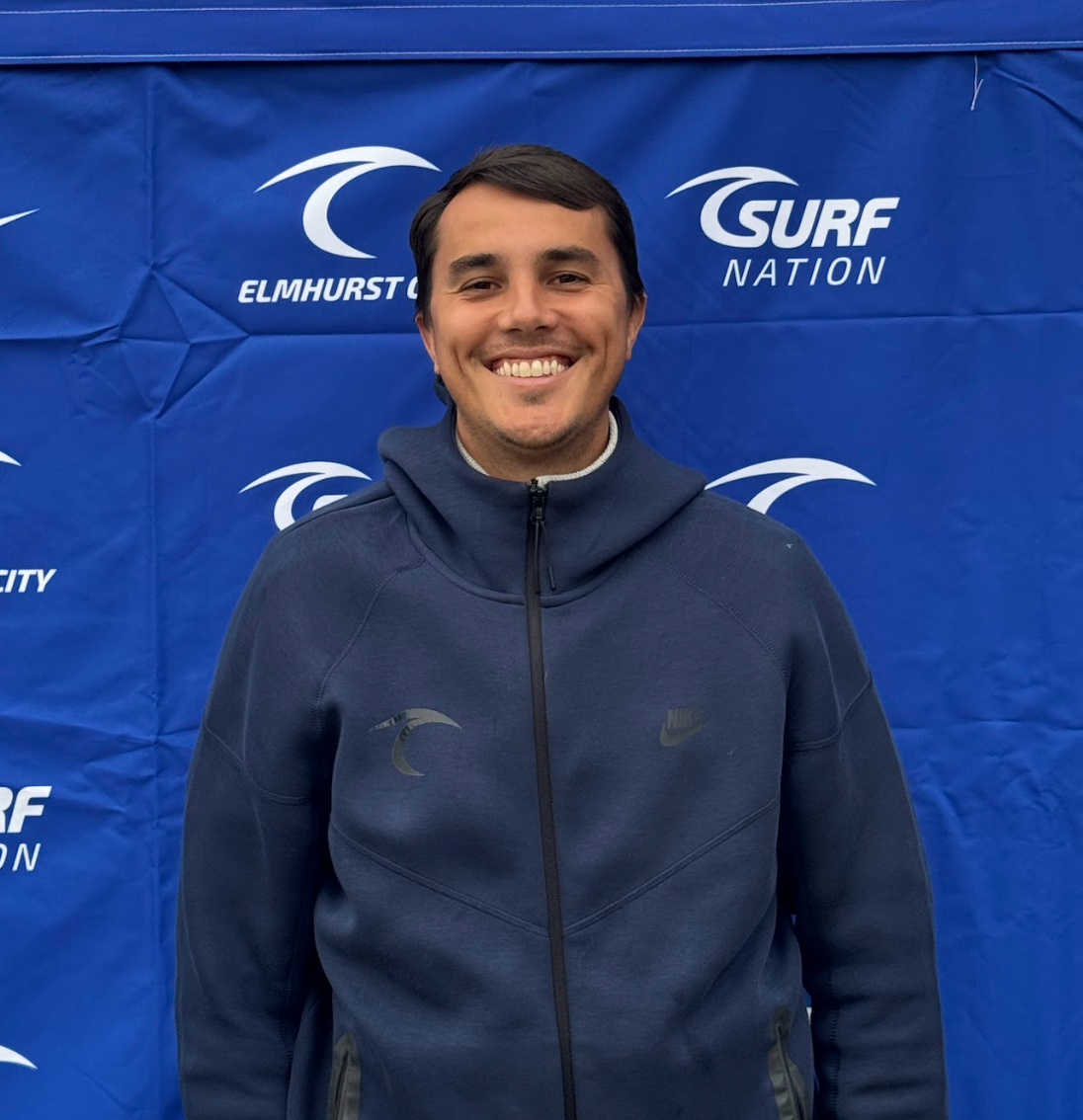A man in a tan shirt is smiling and waving at the camera.
