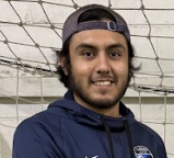 A man wearing a baseball cap and a blue hoodie is standing in front of a soccer net.
