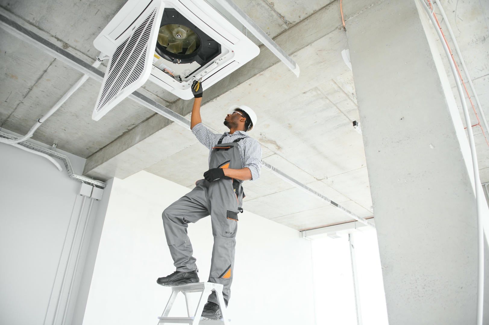 A man is standing on a ladder fixing an air conditioner.