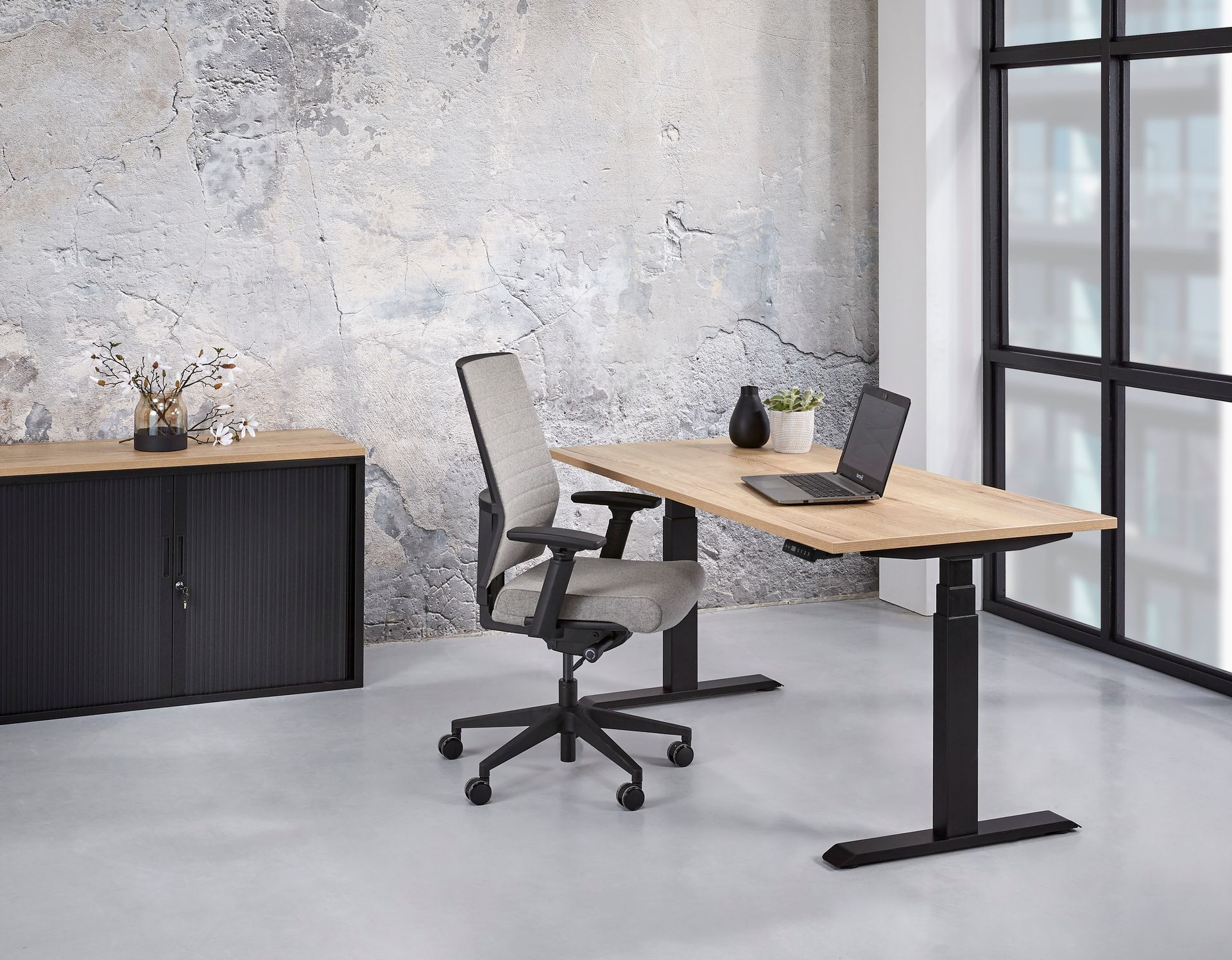 Büro mit einem hellen Holzschreibtisch und einem schwarzen, höhenverstellbaren Untergestell. Am Schreibtisch steht ein grauer Bürostuhl.