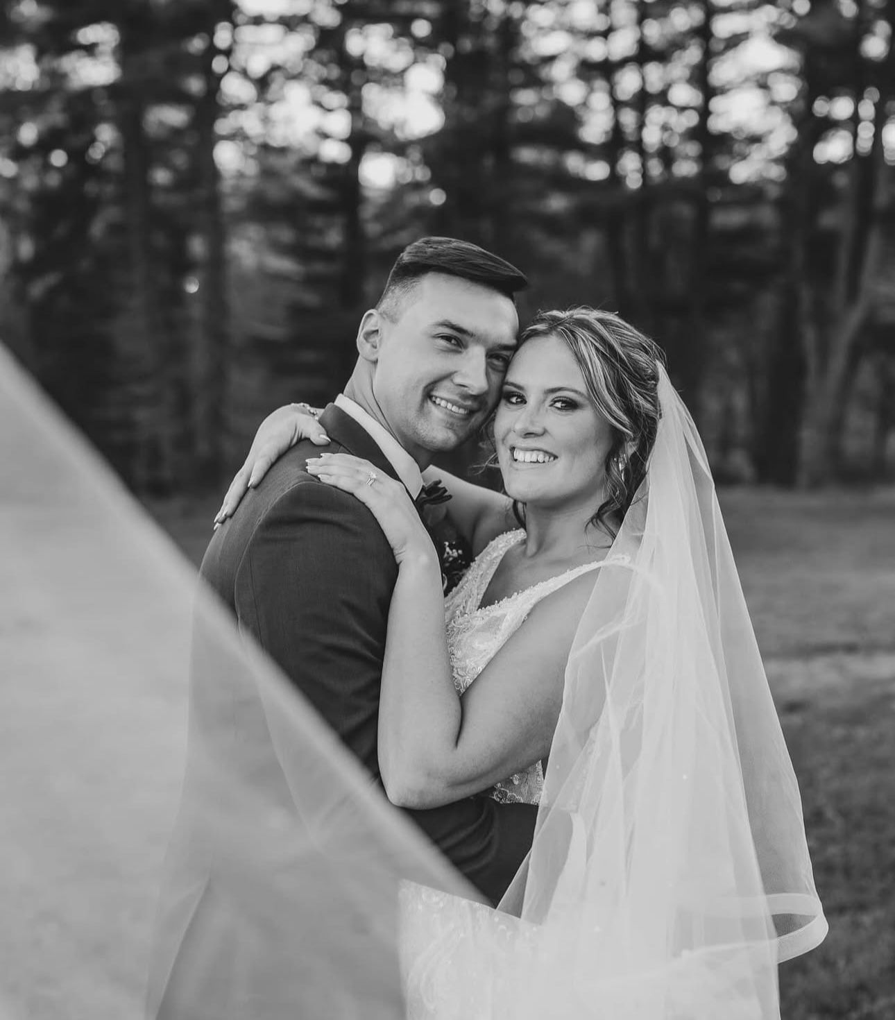 A bride and groom are posing for a black and white photo with their veil blowing in the wind.