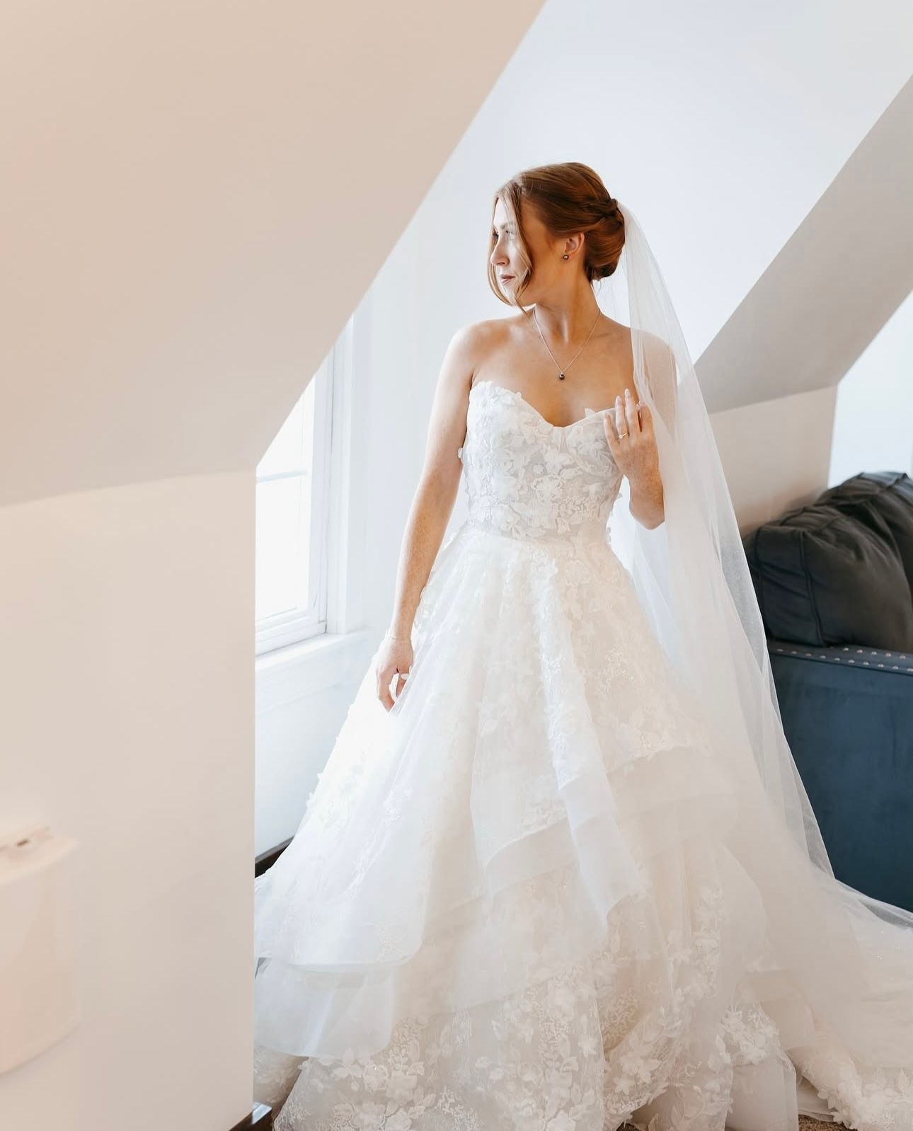 A bride in a white wedding dress and veil is standing in front of a window.