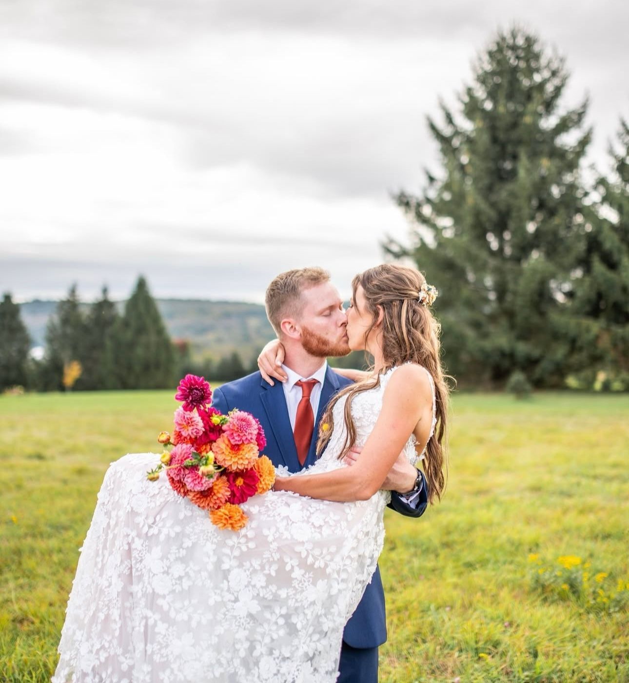 A bride and groom are kissing in a field while the bride is being carried by the groom.