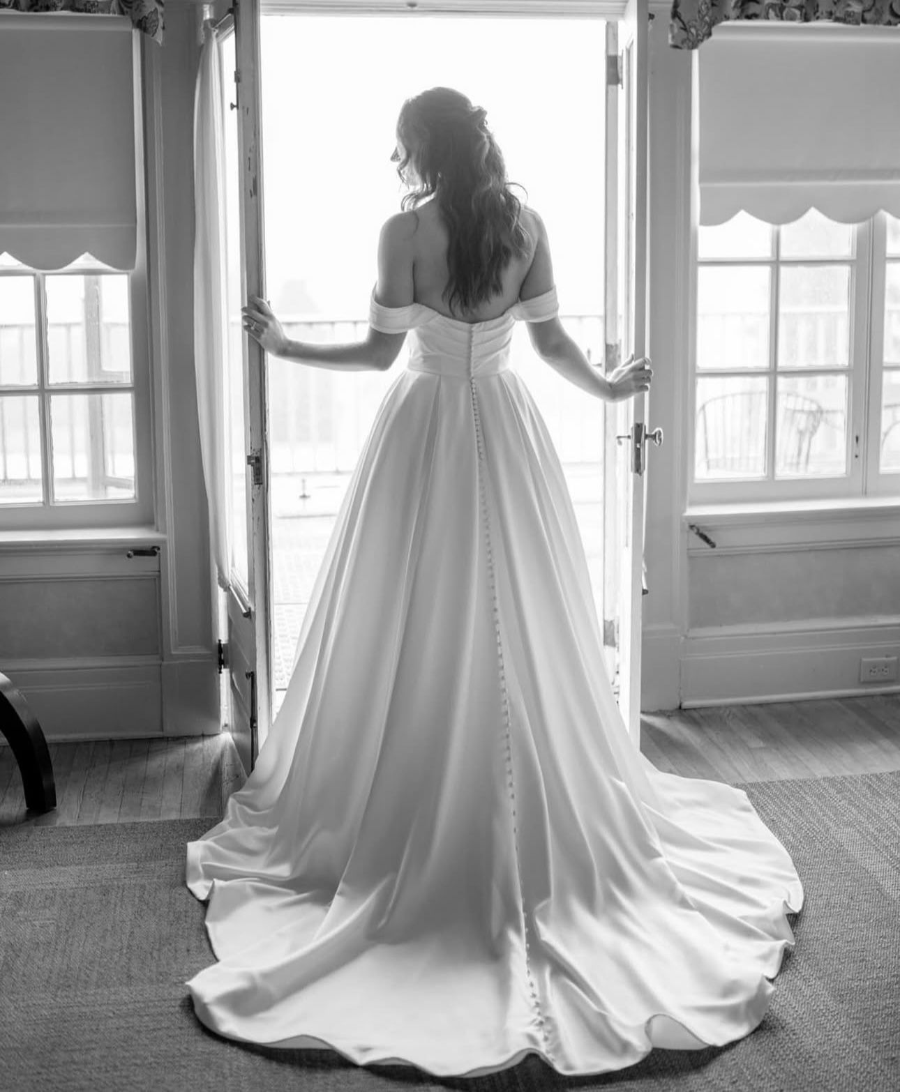 A black and white photo of a bride in a wedding dress standing in front of a window.