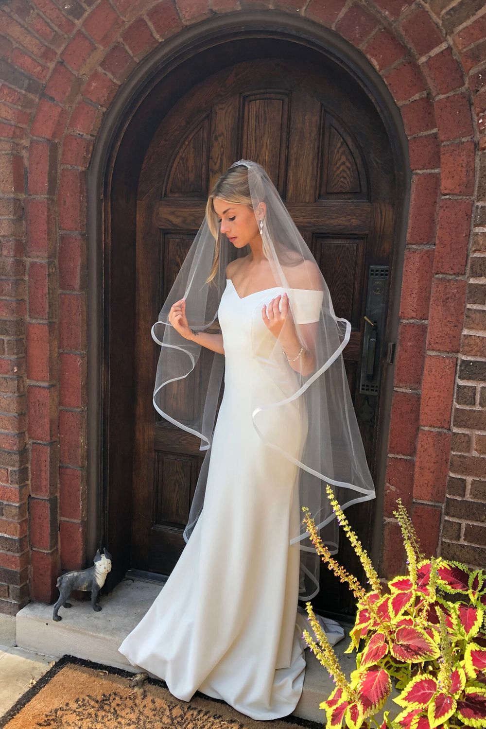A woman in a wedding dress and veil is standing in front of a brick door.