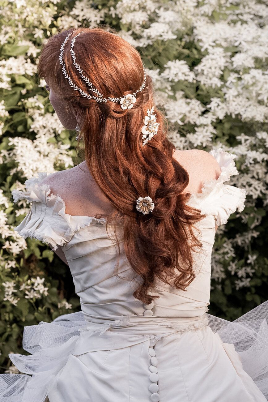 A woman in a white dress with red hair is standing in front of a bush of white flowers.
