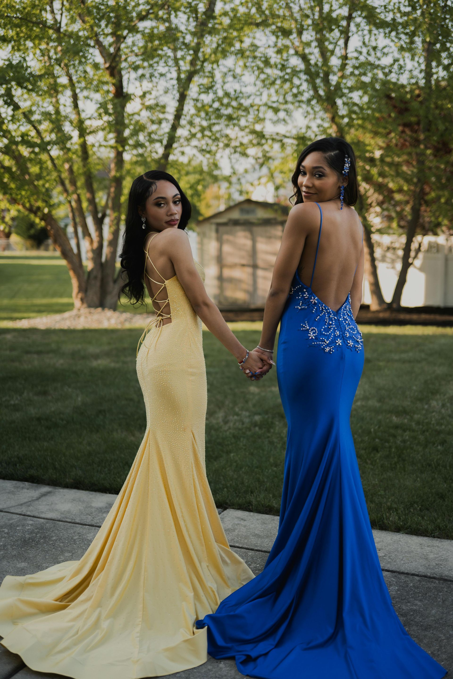 Two women in prom dresses are standing next to each other holding hands.