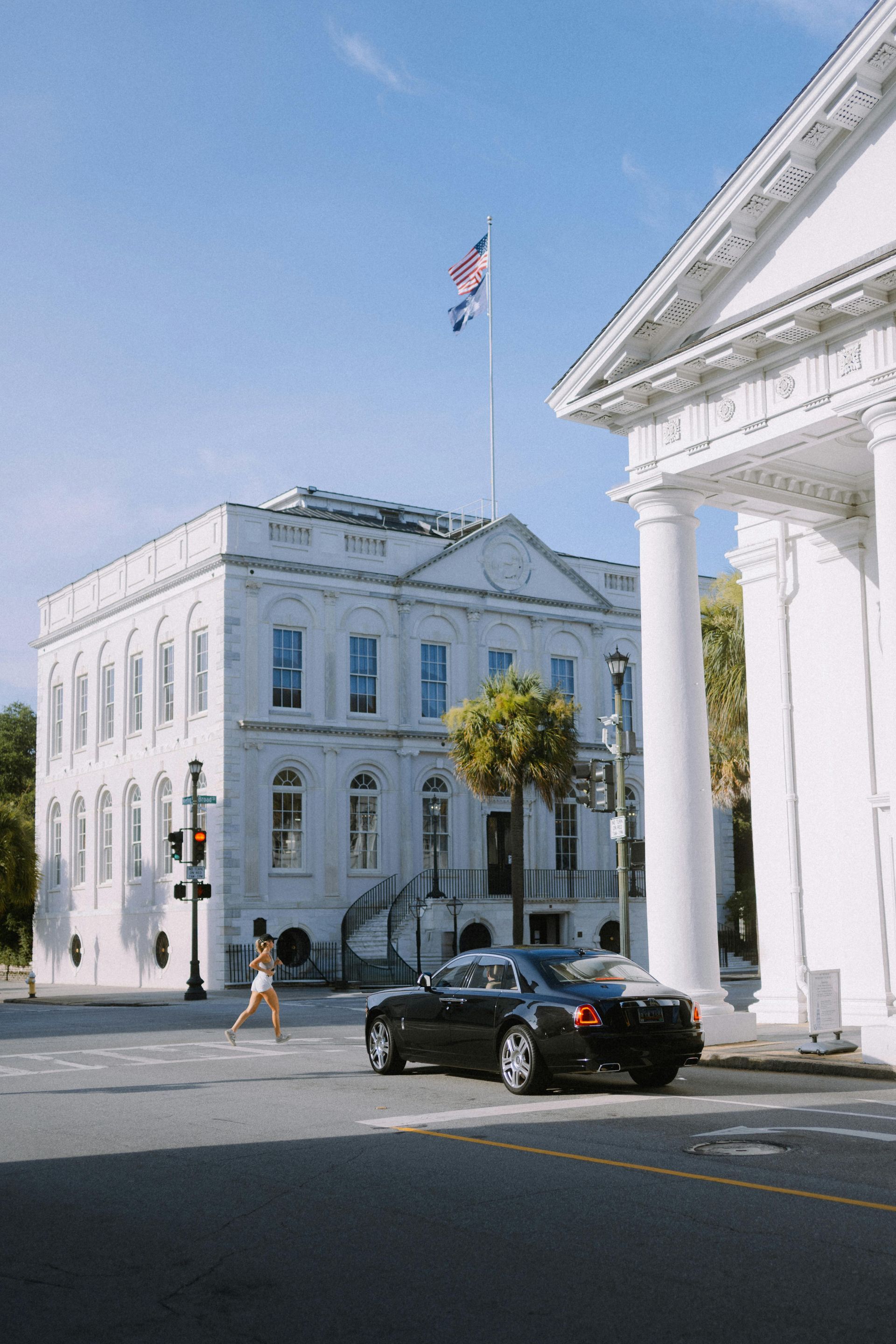 White buildings with columns, flag, car, person crossing street. Sunny day.