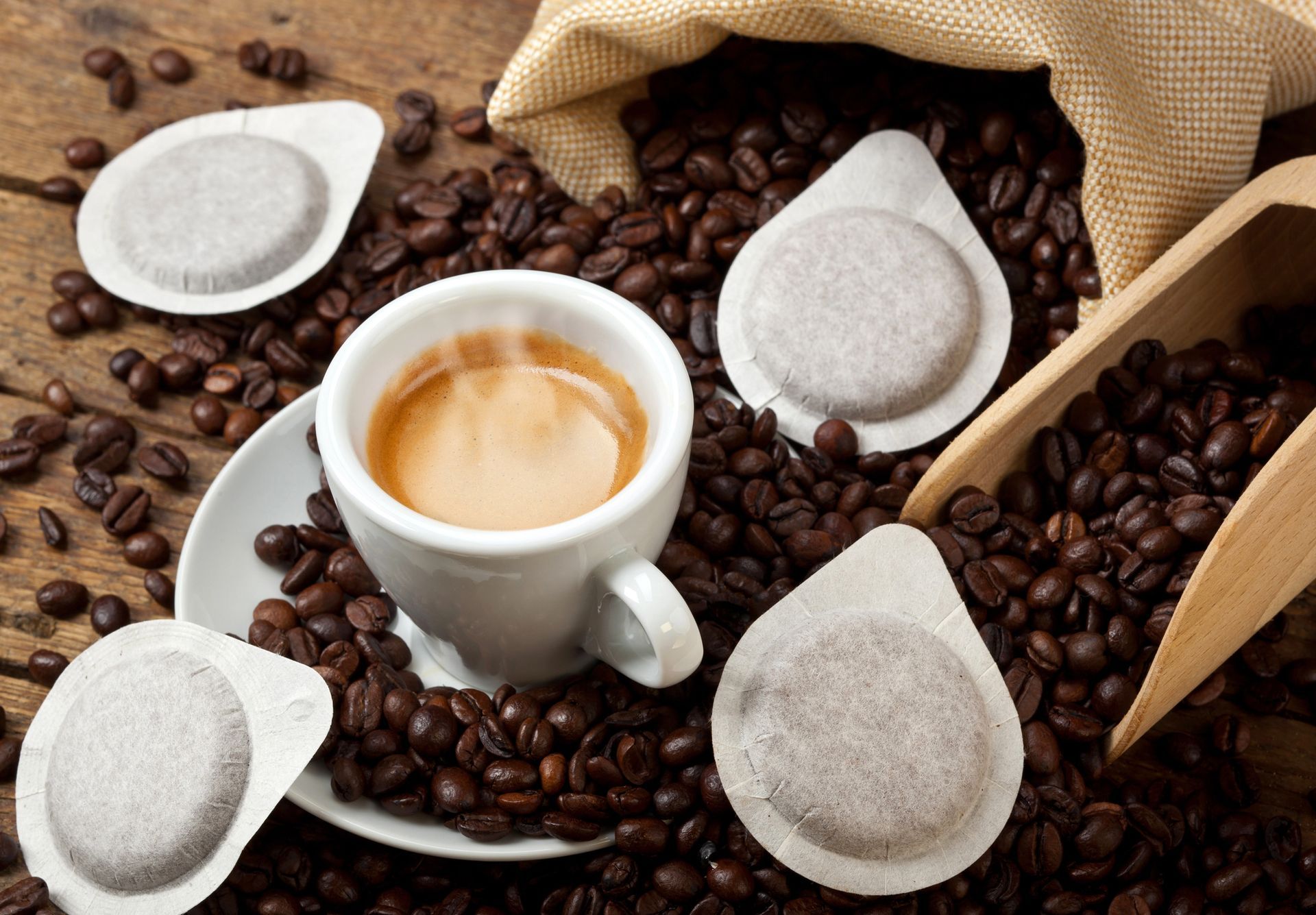 Una tazza bianca di espresso fumante, circondata da chicchi di caffè sfusi e diverse capsule di caffè su una superficie di legno.