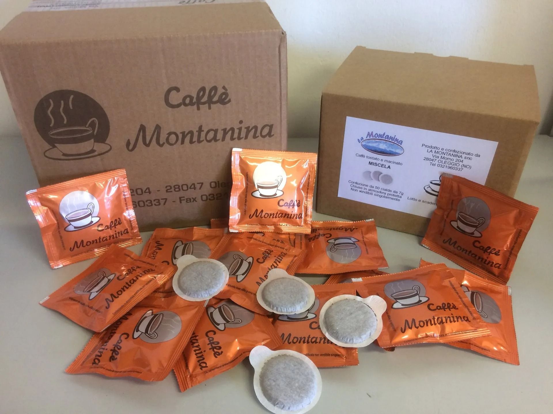 Due scatole di cartone con il marchio Caffè Montanina, circondate da singole capsule di caffè avvolte in carta arancione, su una superficie piana.