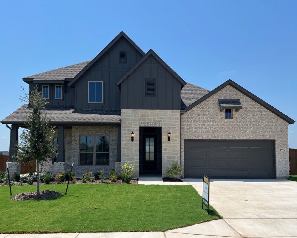 Two-story house with gray roof, stone facade, gray garage door, and lush green lawn on a sunny day.