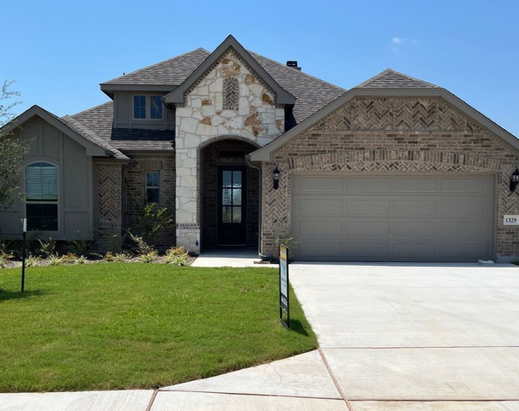 A two-story house with a tan garage door, brick and stone exterior, and green lawn.