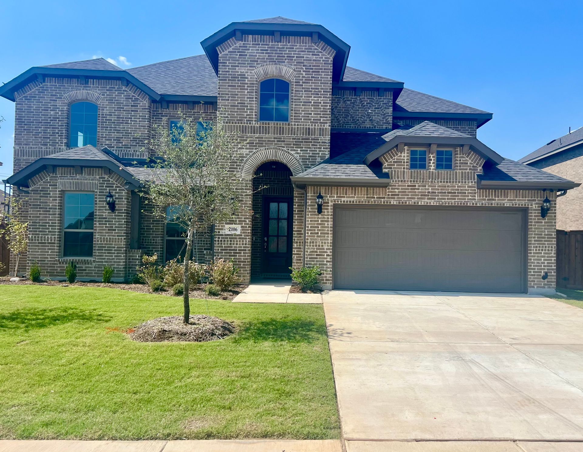 Two-story brick house with stone accents, gray roof, garage door, green lawn, and blue sky.