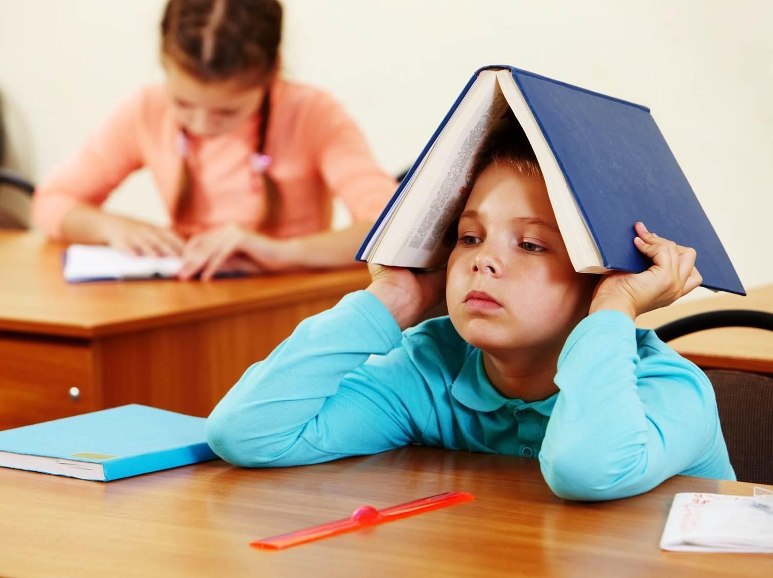 Criança sentada em uma cadeira escolar com um livro sobre a cabeça e com expressão de tédio.