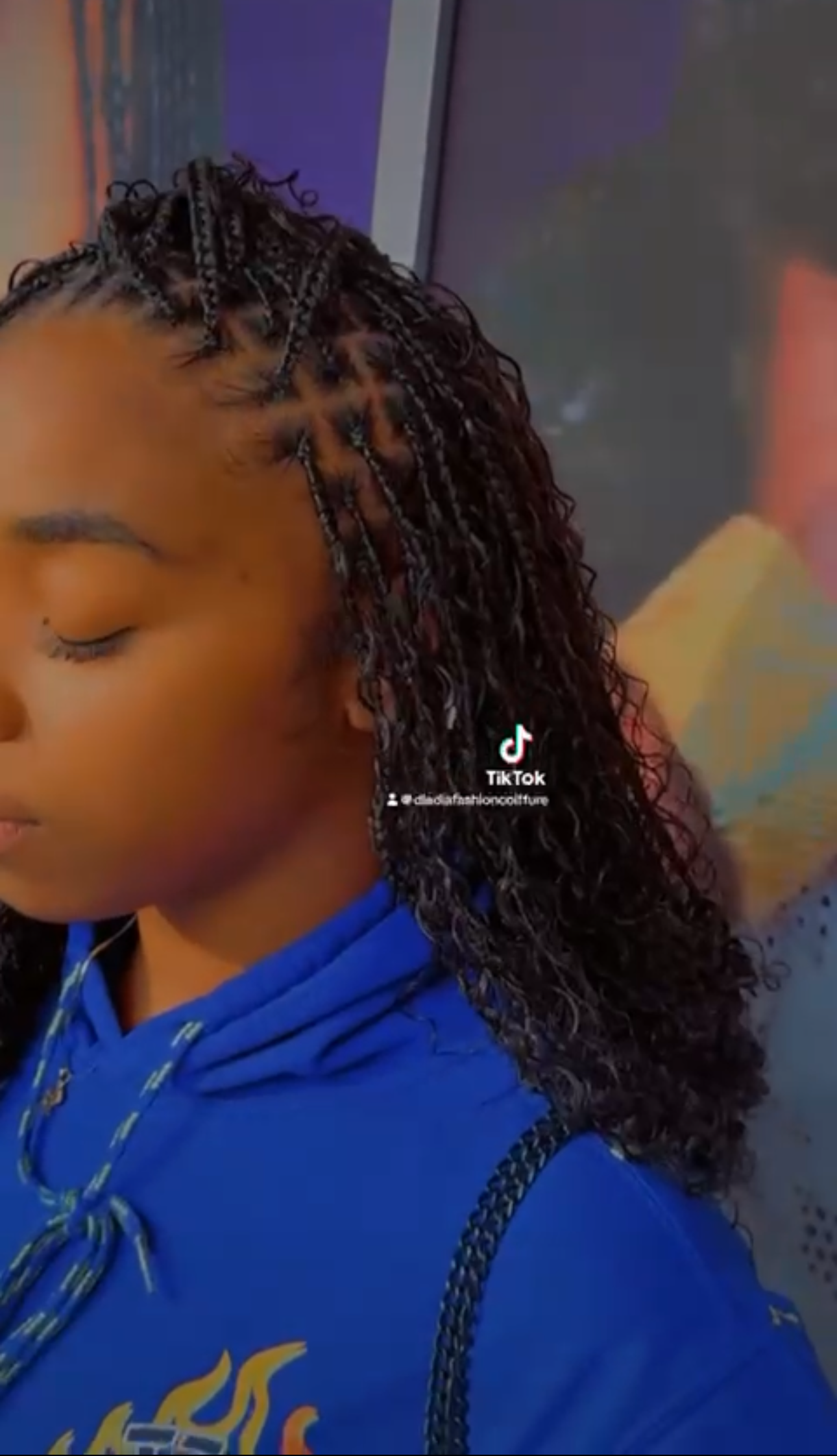 Woman with micro braids, some styled into a geometric design, wearing a blue hoodie.