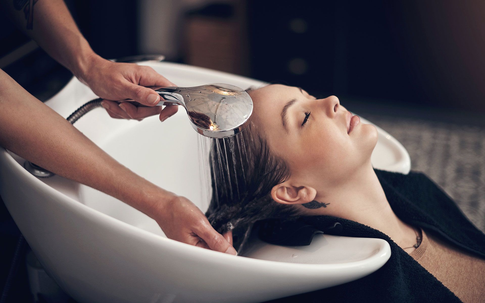Woman getting hair washed in a salon sink. Hairdresser holds showerhead, water streaming on hair.