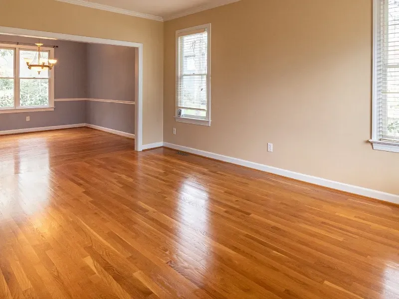 Empty room with hardwood floors, beige walls, and windows; a doorway leads to a gray-walled dining area.