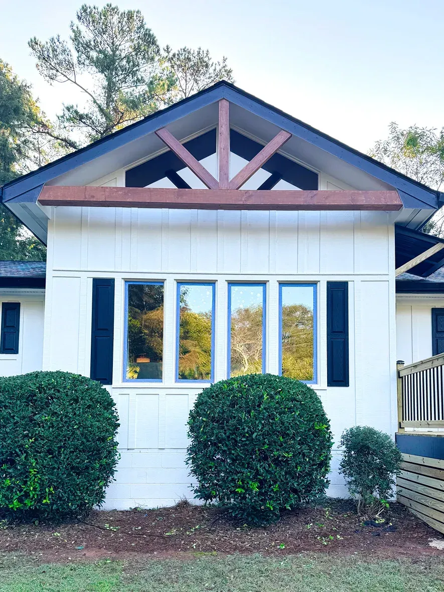 White house with dark trim and triangular wooden detail above windows; green bushes in front.