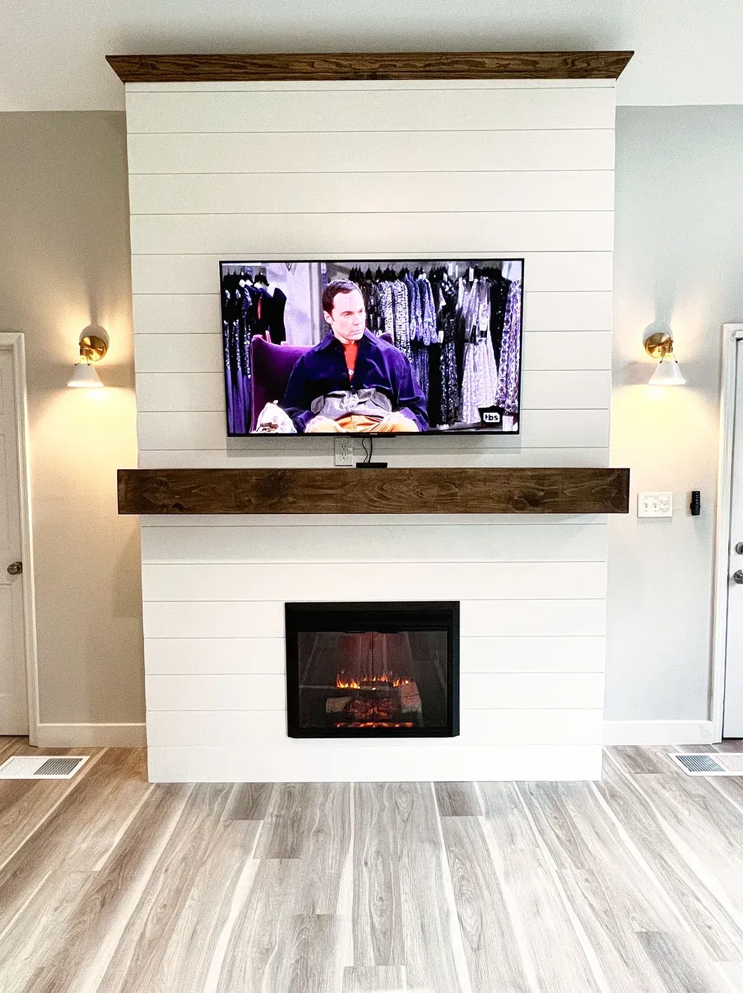 Fireplace with TV mounted above, white shiplap, wood mantel, and sconces. The TV is on.