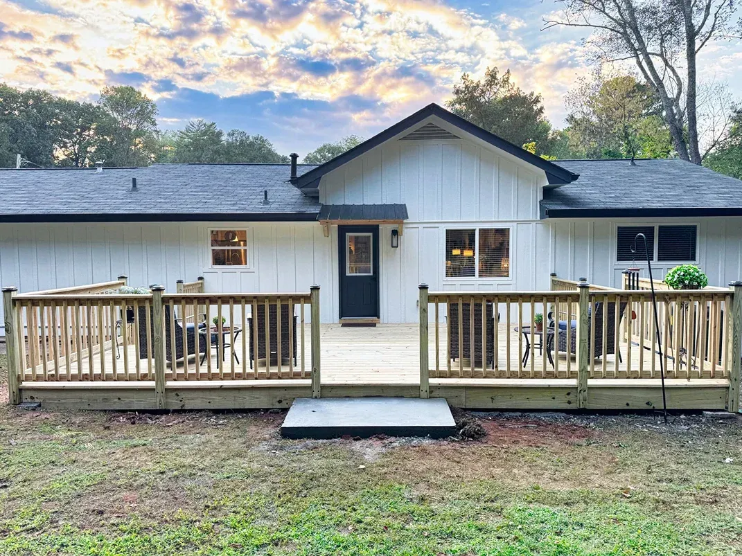 White house with wooden deck and railings in a grassy yard, under a partly cloudy sky.