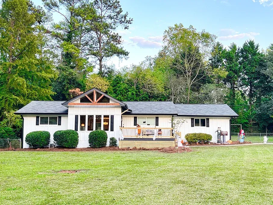 White one-story house with black shutters, wooden accents, and a large front lawn, surrounded by trees.