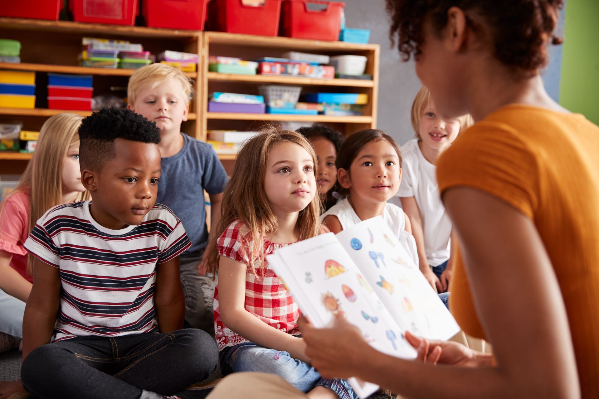 A woman reading a storybook to a group of young children sitting on the floor in a colorful room.