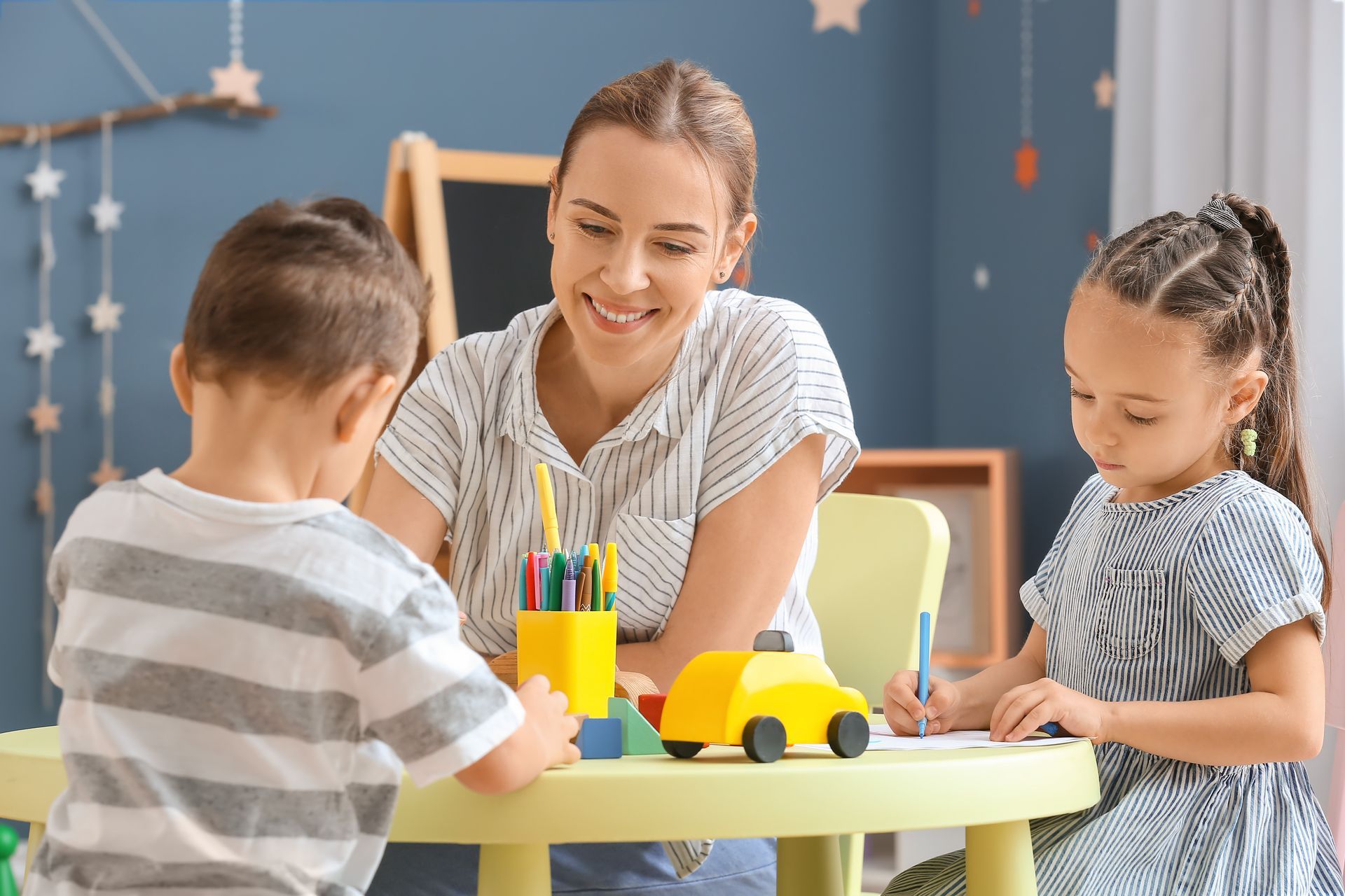 An adult smiles as two children draw and play at a small round table. An adult smiles as two children draw and play at a small round table.