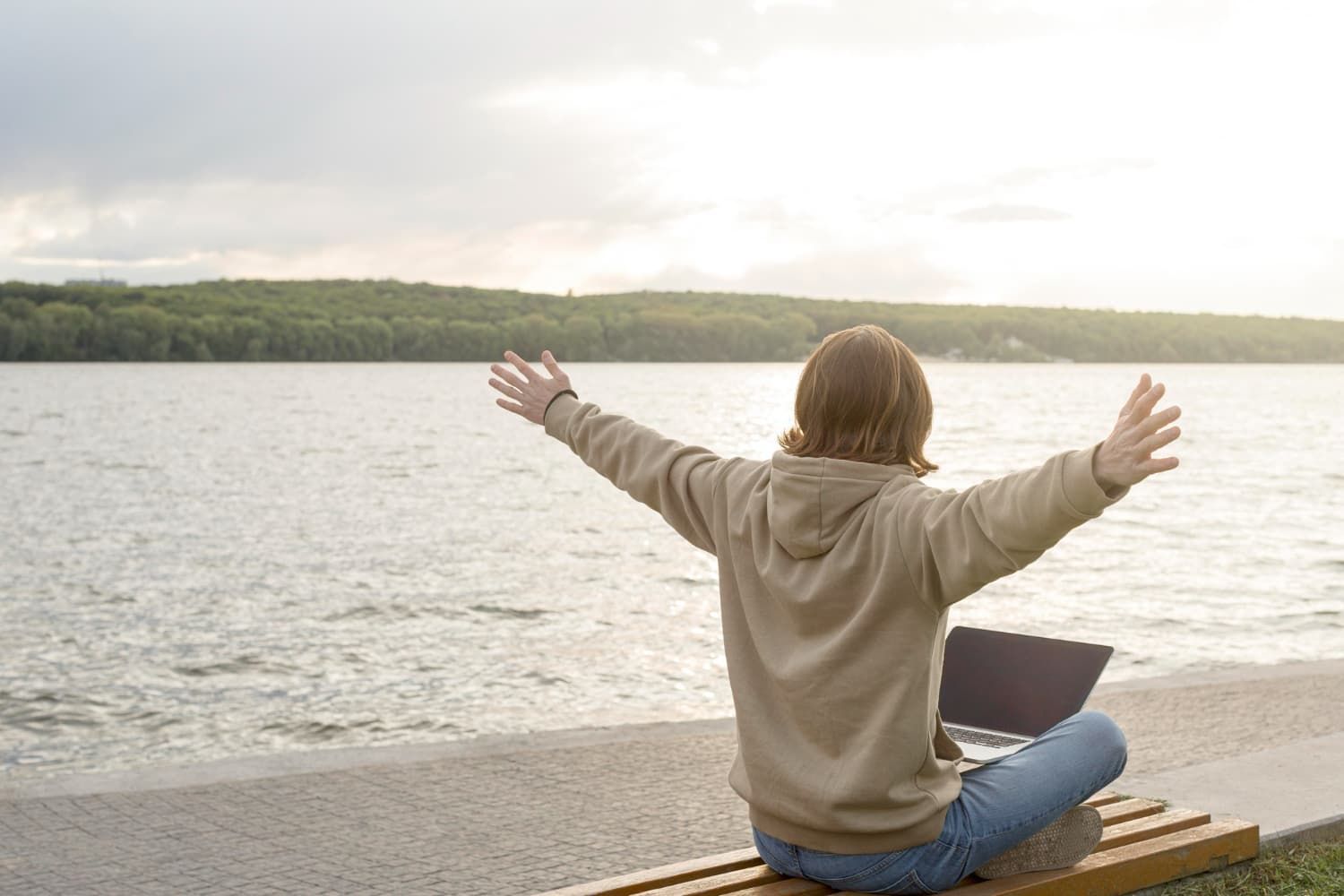 A woman is sitting on a bench with her arms outstretched in front of a lake.