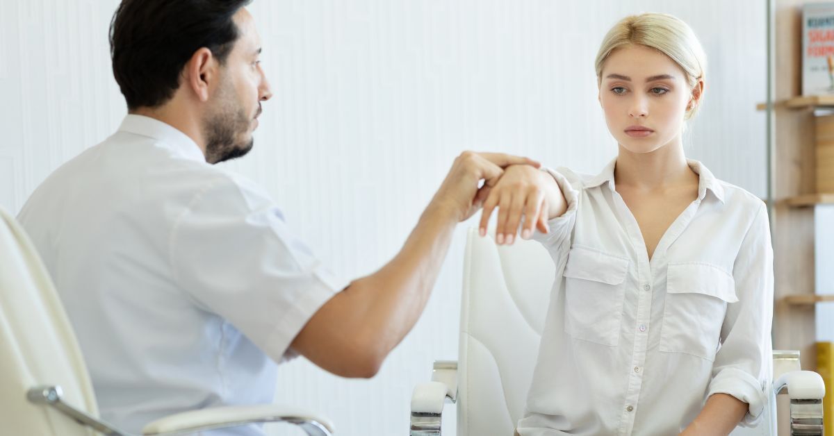 A man is examining a woman's arm while she sits in a chair.