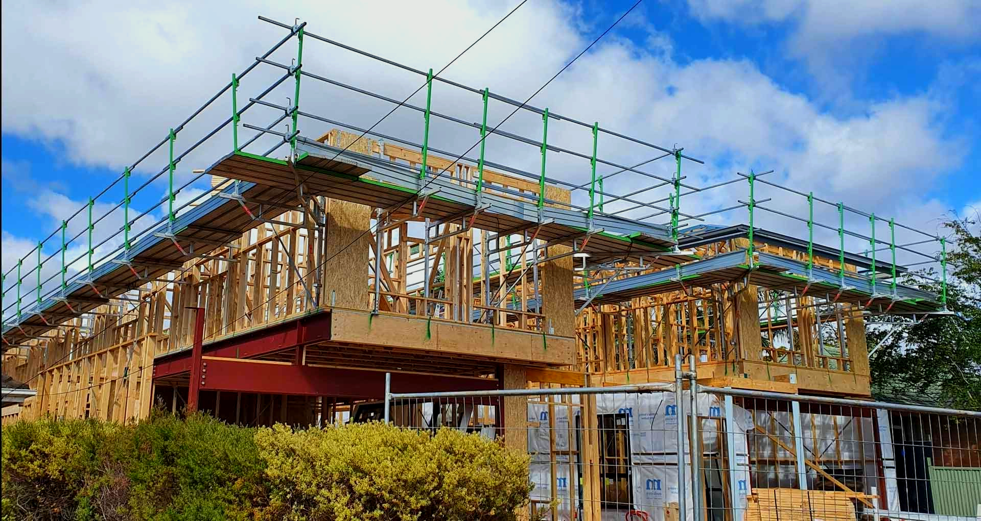 Construction site: wood-framed building under scaffolding, green rails. Cloudy blue sky.