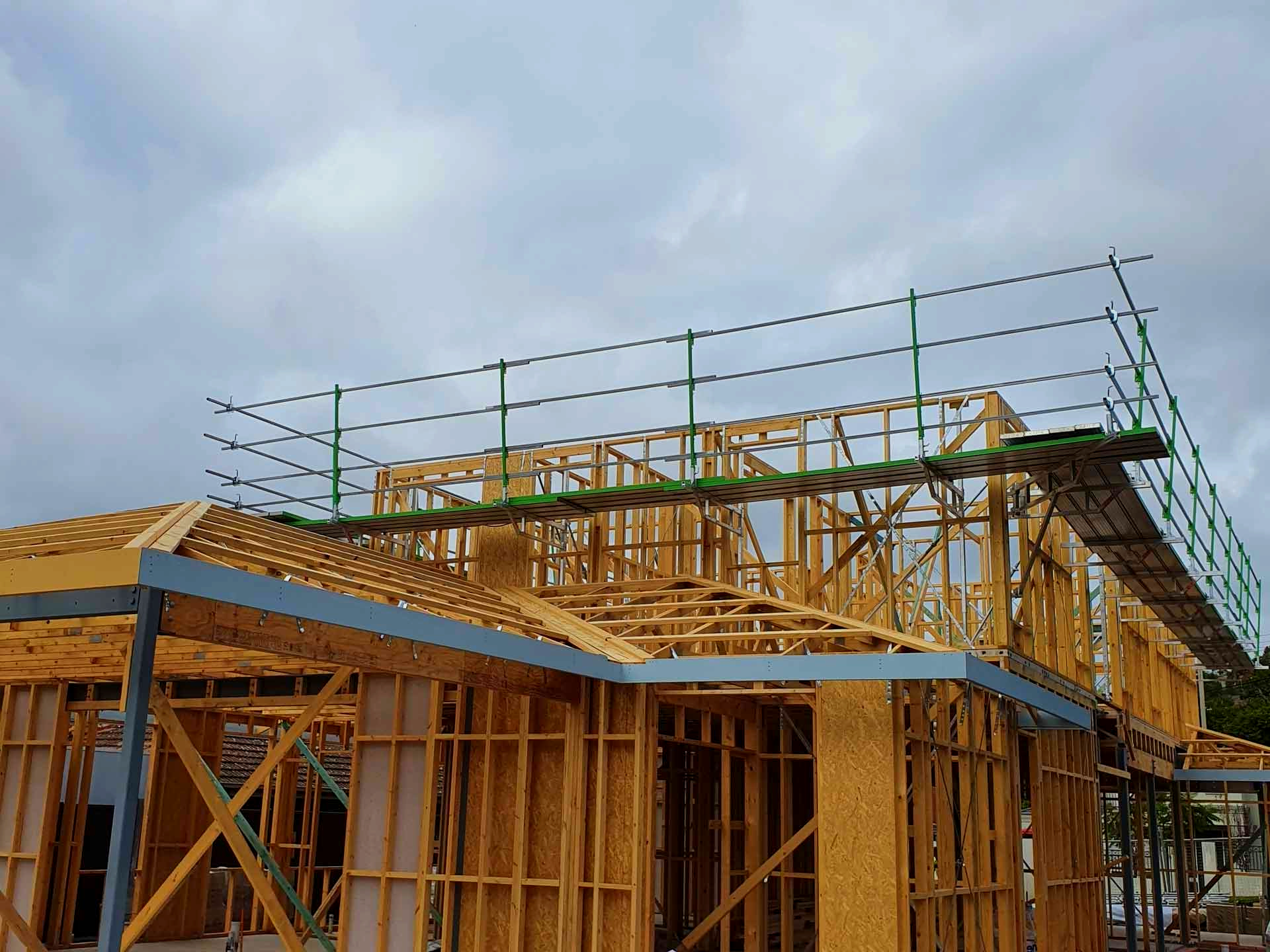 Wooden house frame under construction with green safety rails on the roof. Overcast sky.