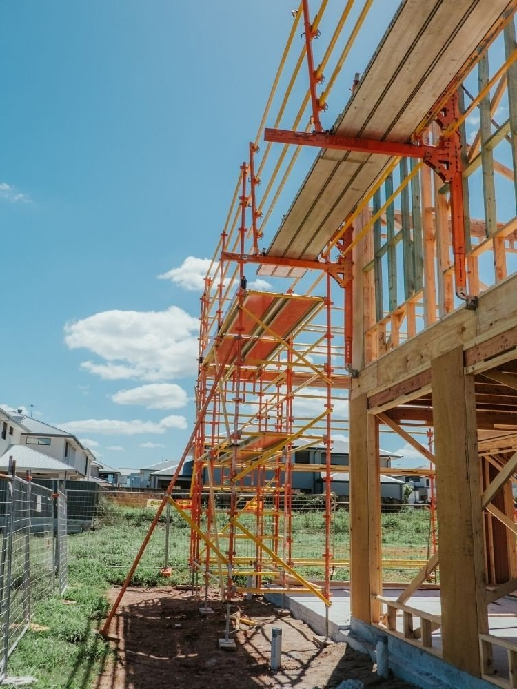 Construction scaffolding next to a wooden house frame under a blue sky.