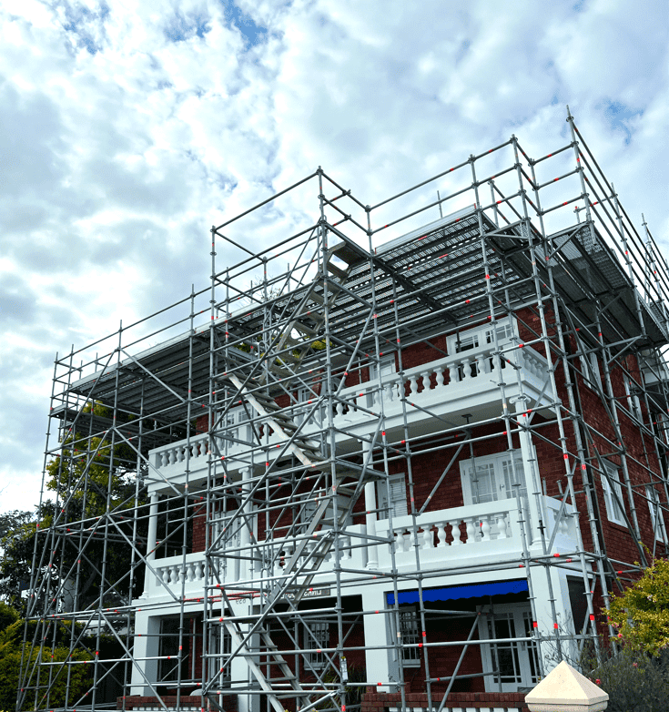 Scaffolding surrounds a red brick building with white balconies. Cloudy sky in the background.