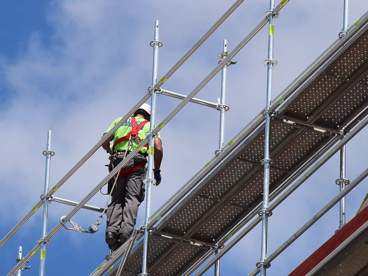 Construction worker on scaffolding, wearing safety harness, against a blue sky.