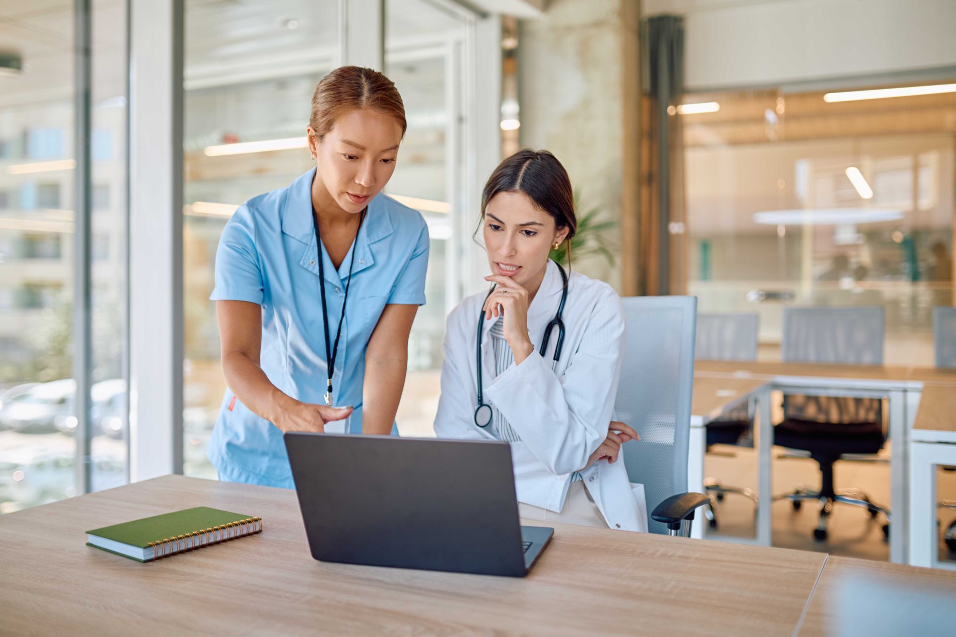 Two healthcare workers reviewing laptop at a desk. One points, the other ponders. Modern office.