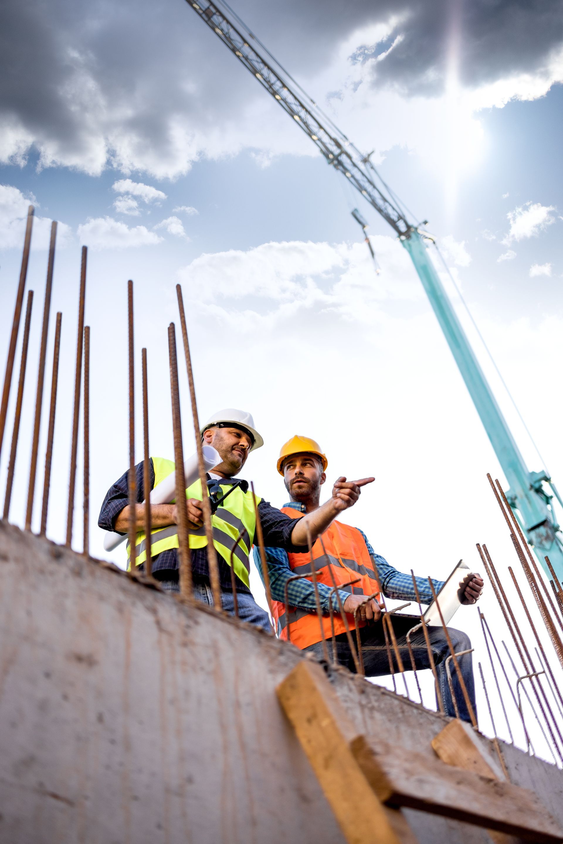 Deux ouvriers du bâtiment sur une structure en béton ; l'un montre du doigt, l'autre tient des plans, une grue en arrière-plan, ciel ensoleillé.