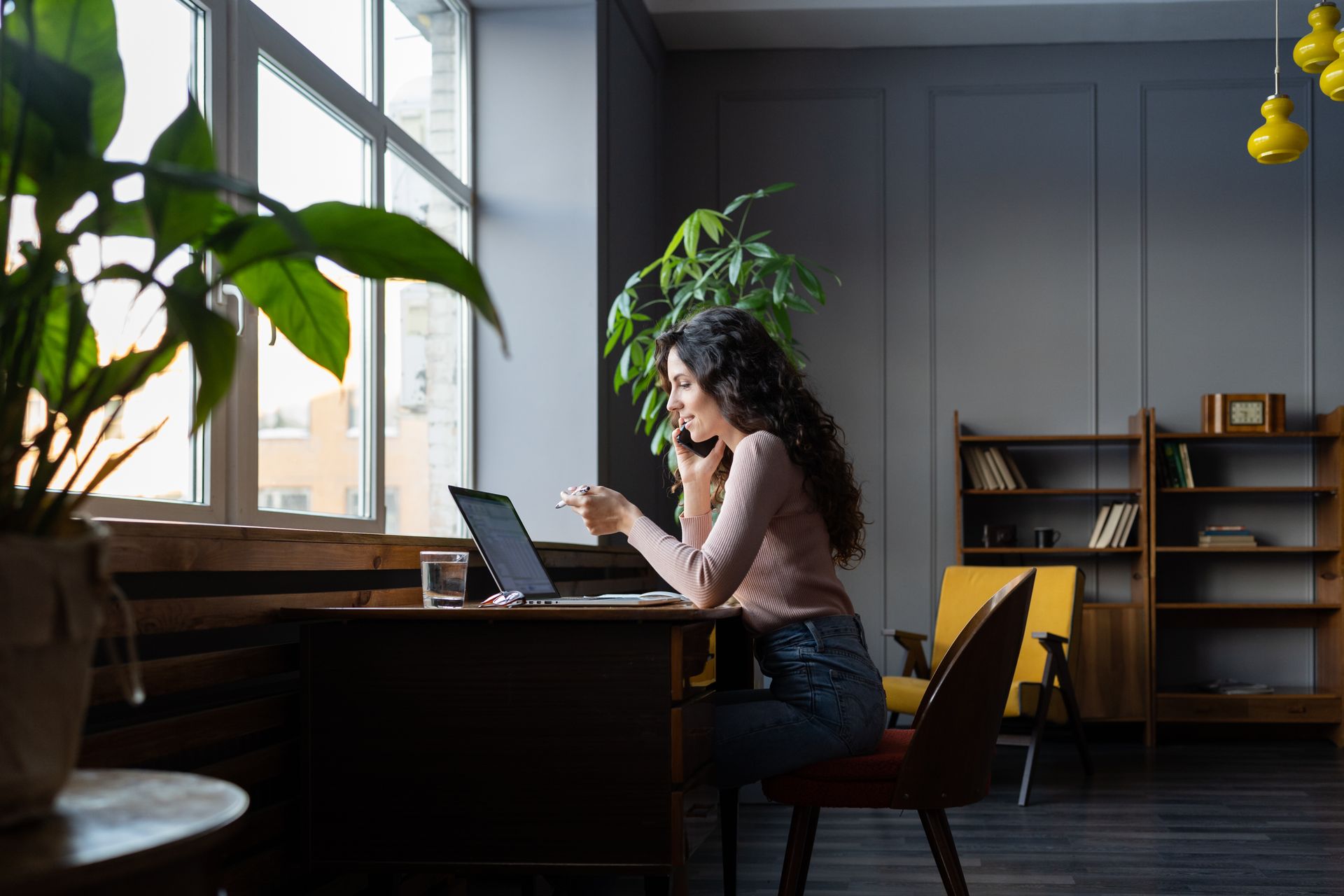 A woman is sitting at a desk with a laptop and talking on a cell phone.